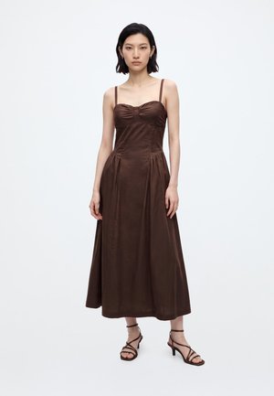 Woman with short dark hair wearing a sleeveless brown midi dress and brown strappy heeled sandals, standing against a plain white background.