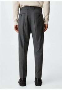 Personne portant un pantalon taille haute gris foncé sur mesure avec des plis et des poches arrière boutonnées, des chaussures noires et un pull beige.