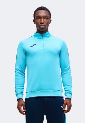 Homme portant un haut de sport bleu clair à manches longues avec fermeture éclair au quart et pantalon bleu foncé, debout devant un fond uni et clair.