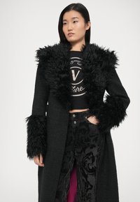 Črni plašč z teksturirano tkanino, ki ima voljen umetni krzneni ovratnik in manšete. Nošen čez črno majico in temne hlače z vzorcem.