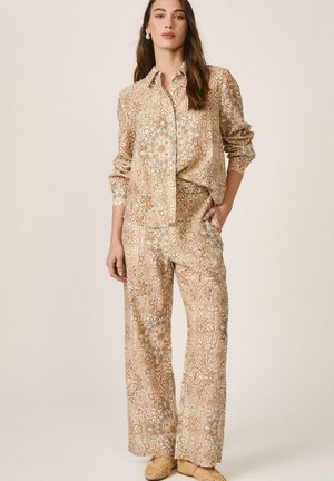 Mujer vestida con camisa beige de manga larga con botones y patrón, y pantalones anchos a juego, con zapatos tipo slip-on tejidos, frente a un fondo liso.