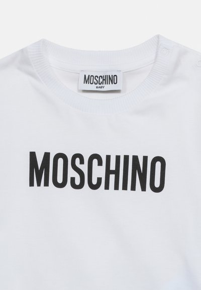 Λευκό παιδικό μπλουζάκι με μαύρο κείμενο "MOSCHINO" στο μπροστινό μέρος και ταυτόχρονες ετικέτες στο εσωτερικό του γιακά, με κουμπιά πατιλέτας στον έναν ώμο.