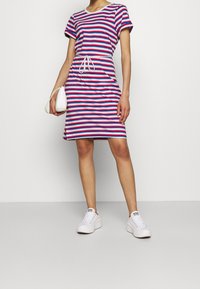 Robe rayée avec des bandes horizontales rouges, bleues et blanches, manches courtes, taille à cordon de serrage, fentes latérales, associée à des baskets blanches.