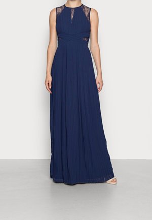 Granatowa sukienka maxi bez rękawów, plisowana, z koronkowymi detalami na ramionach i górnej części dekoltu, noszona z beżowymi butami na otwartym palcu.