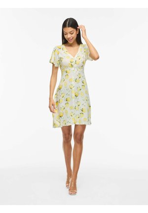 Mujer con vestido blanco de manga corta con estampado de limones amarillos, caminando, tocándose el pelo y llevando tacones con tiras transparentes sobre un fondo blanco.