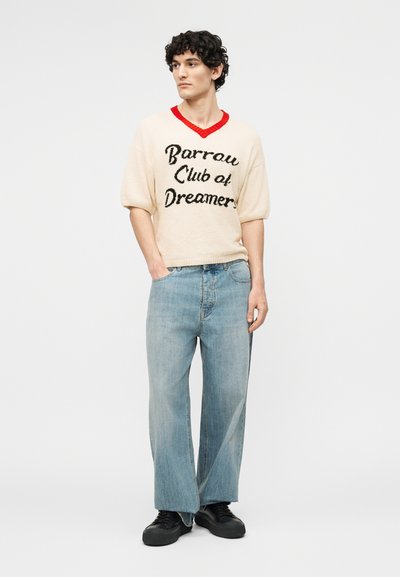 Jeune homme portant un pull crème avec col rouge et l'inscription « Barrow Club of Dreamers », jean large clair, et chaussures noires, debout les mains dans les poches.