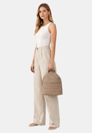 Vrouw in een wit mouwloos shirt en beige broek met rechte pijpen, die een beige rugzak met patroon bij het handvat vasthoudt, staand op een witte achtergrond.