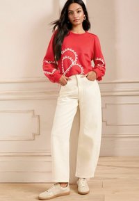 Felpa rossa con motivo decorativo bianco sulle maniche, abbinata a pantaloni cream a vita alta e sneaker beige, su uno sfondo chiaro.