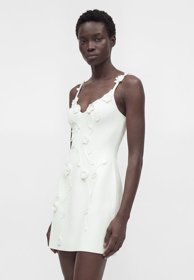 Γυναίκα με λευκό αμάνικο φόρεμα με 3D floral εφαρμογές και λεπτές τιράντες, στέκεται μπροστά σε ένα απλό φωτεινό φόντο.