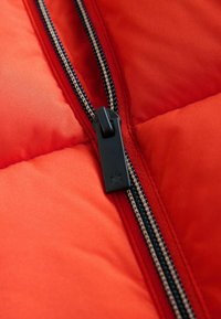 Tissu de doudoune rouge avec gros plan sur une fermeture éclair noire et une tirette. La veste présente des sections matelassées et des coutures contrastées.