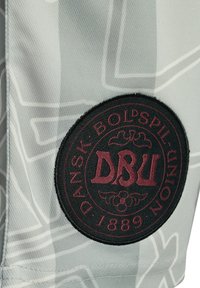 Grått tyg med ett subtilt texturerat mönster och en rund svart lapp som visar "DBU" i vinrött, omgiven av "DANSK BOLDSPIL UNION 1889."