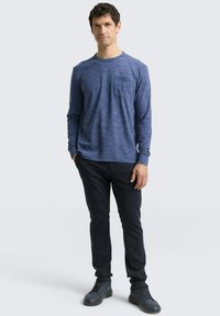 Langärmliges, blaustichiges Hemd mit einer Brusttasche, kombiniert mit dunklen Hosen und schwarzen Turnschuhen. Einfaches Design mit Rundhalsausschnitt.