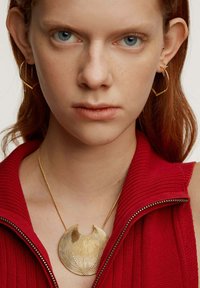 Collar de metal dorado con líneas onduladas y una forma de media luna. Modelo lleva una prenda de punto roja con cremallera. Pendientes de aro geométricos dorados.