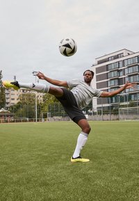 Atleta con una camiseta gris y pantalones cortos negros, calcetas a rayas blancas, realizando una patada con un balón de fútbol blanco y negro en un campo verde.