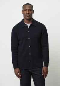 Cárdigan de punto acanalado en navy con botones, collar plano, mangas largas y botones oscuros, combinado con una camisa gris debajo.