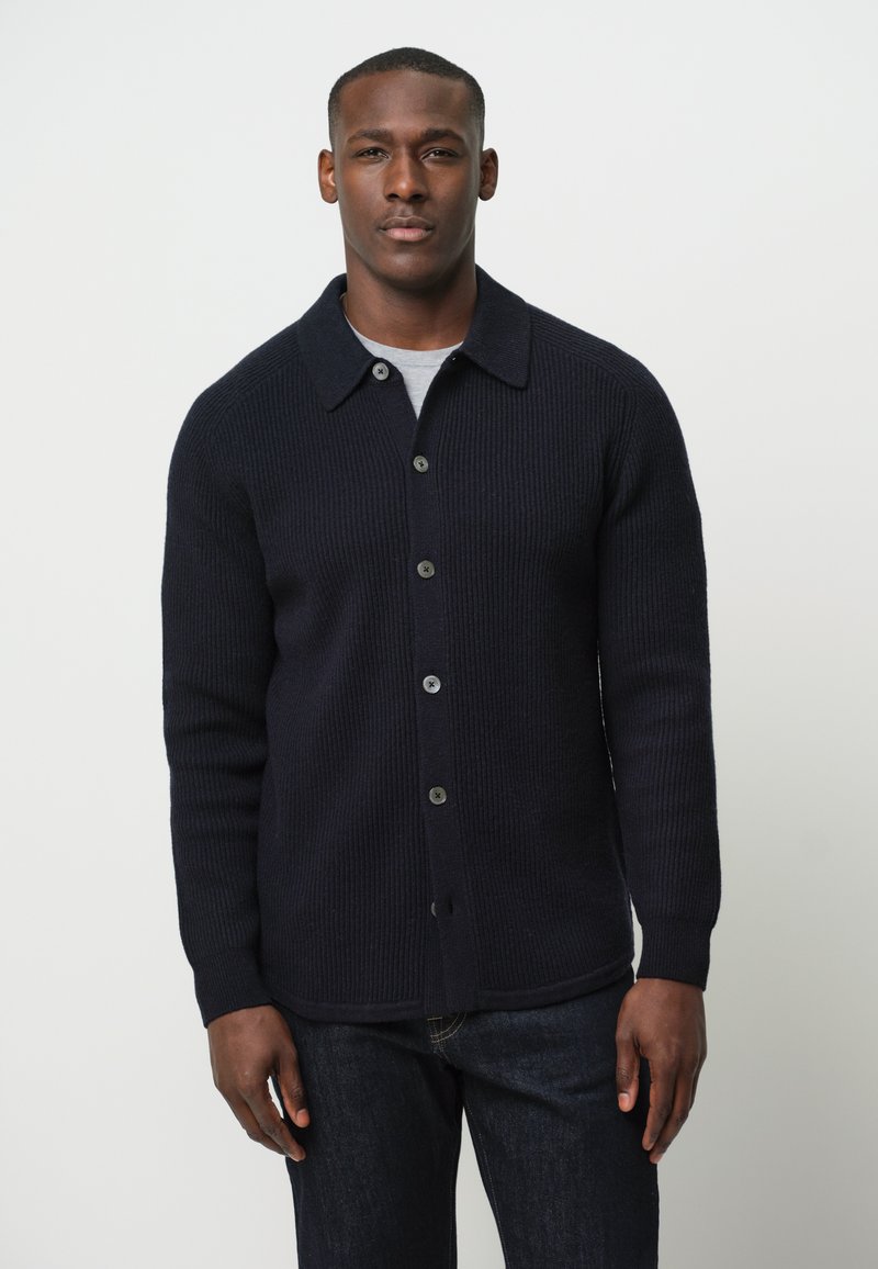 Cárdigan de punto acanalado en navy con botones, collar plano, mangas largas y botones oscuros, combinado con una camisa gris debajo.