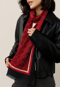 Red scarf featuring a black abstract pattern, with a contrasting border in red and beige, wrapped loosely around the neck.