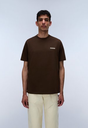 Homme portant un t-shirt marron foncé à manches courtes avec un petit logo blanc sur la poitrine et un pantalon beige clair, debout devant un fond uni.