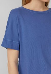 Niebieska bawełniana koszulka z okrągłym dekoltem, krótkimi rękawami i widocznymi szwami wzdłuż dołu i brzegów rękawów. Gładka faktura.