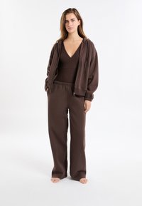 Brązowy zestaw do wypoczynku, składający się z bluzy z kapturem zapinanej na suwak, topu z głębokim dekoltem w serek oraz luźnych spodni z elastycznym pasem, wszystko wykonane z miękkiego materiału.