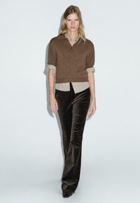 Bruin gebreid sweater met korte mouwen over een licht button-up shirt, gecombineerd met donkere, flared fluwelen broek en zwarte schoenen.