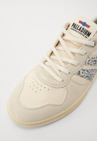 Baskets beiges avec un design en nubuck et en maille ; coque de protection perforée ; accent avec imprimé animal bleu et gris. Les lacets sont de couleur crème.