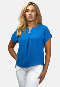 Niebieska bluzka krótki rękaw z dekoltem w serek, teksturowany materiał oraz detal w postaci guzika przy mankiecie. Zestawiona z dopasowanymi białymi spodniami.