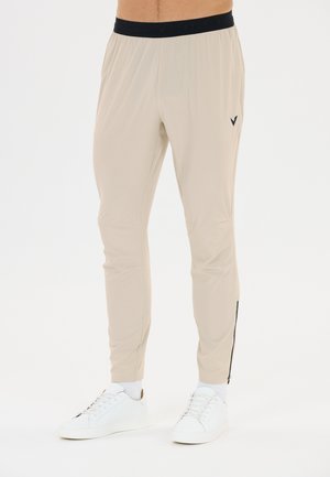 Beige Slim-Fit-Jogginghose mit schwarzem elastischem Bund und seitlichen Reißverschluss-Details, getragen mit weißen Sneakers und Socken.