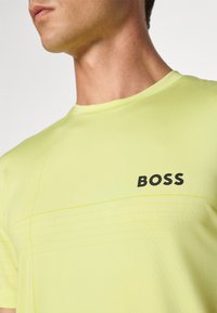 Gul atletisk t-skjorte med rund hals. Har et vertikalt teksturert mønster og en svart "BOSS" logo på brystet.