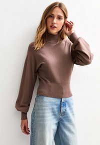 Vrouw in een bruine coltrui met ballonmouwen en hoogwaisted lichtblauwe jeans, poserend tegen een witte muur.