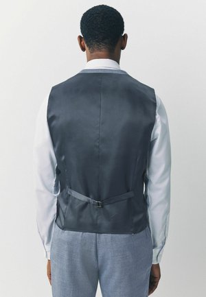 Chaleco de satén gris con una textura suave, que presenta una correa y una hebilla en la parte posterior, usado sobre una camisa de color claro.