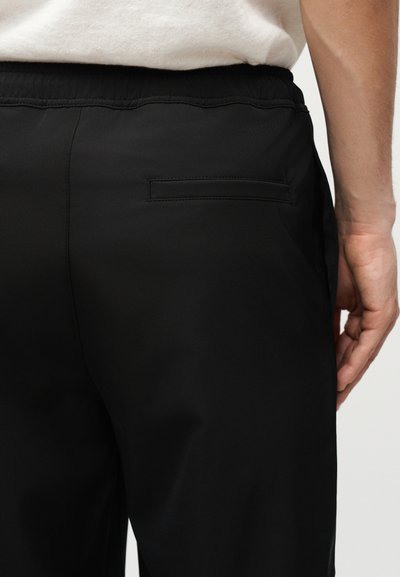 Κοντινό πλάνο ενός ατόμου που φοράει μαύρο παντελόνι με τσέπη στο πίσω μέρος και ελαστική μέση, και λευκό πουκάμισο μπροστά μέσα στο παντελόνι.