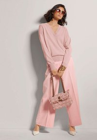 Helles rosa Strickpullover mit V-Ausschnitt, kombiniert mit hellrosa weit geschnittenen Hosen. Accessoires: eine strukturierte rosa Handtasche und weiße Absatzschuhe.