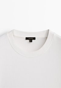 White cotton t-shirt featuring a ribbed crew neckline and a label with brand name and size tag, viewed from above. Smooth texture.