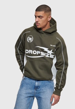 Homme portant un sweat à capuche vert olive avec le texte et le graphisme "DROPSIZE", assorti à un jean bleu clair, regardant vers le bas à sa gauche.