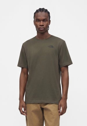 Ung mand med fletninger iført en olivengrøn The North Face t-shirt og beige bukser, der står mod en ensfarvet hvid baggrund.