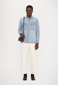 Cămașă din denim bleu deschis cu buzunare pe piept, pantaloni largi de culoare crem și mocasini negri. O geantă maronie este purtată pe umăr.