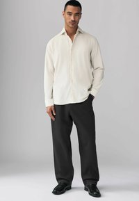 Chemise crème à boutons, à manches longues, associée à un pantalon noir ample et des chaussures noires, présentée sur un fond neutre.