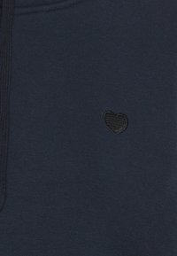 Tissu bleu foncé avec un petit cœur noir brodé sur le côté droit et une couture texturée le long du bord.