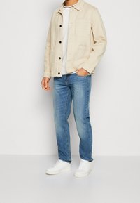 Chaqueta beige de algodón con cierre de botones y bolsillos delanteros, camiseta blanca de cuello redondo, vaqueros azul claro y zapatillas blancas.