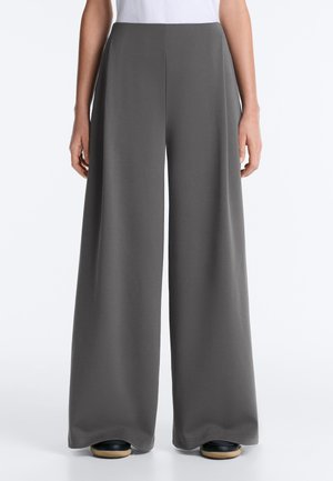 Osoba ubrana w ciemnoszare, szerokie spodnie z wysokim stanem, połączone z czarnymi butami i białą bluzką, stoi na jednolitym tle.