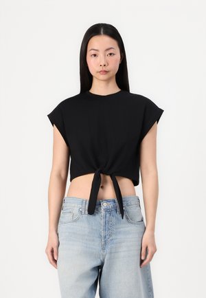 Schwarzes, kurz geschnittenes T-Shirt aus weichem Stoff, mit kurzen Ärmeln und einem Knoten-Detail vorne. Kombiniert mit hellblauen, hochtaillierten Jeans.