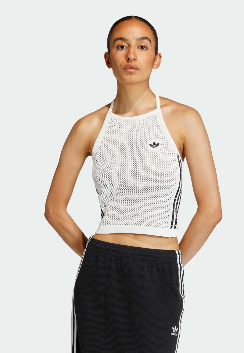 Mladá žena v bílém síťovaném halter topu a černých kalhotách Adidas s bílými pruhy, stojící se založenýma rukama za zády na jednobarevném pozadí.