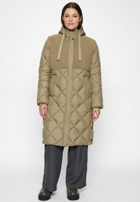 Woman wearing a long quilted beige coat with fleece chest panel and hood, grey wide-leg pants, and dark platform boots, standing straight.