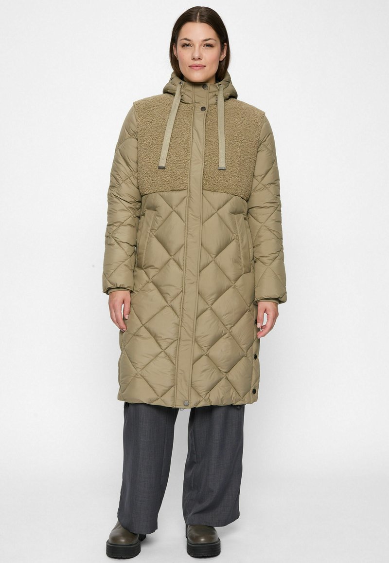 Woman wearing a long quilted beige coat with fleece chest panel and hood, grey wide-leg pants, and dark platform boots, standing straight.