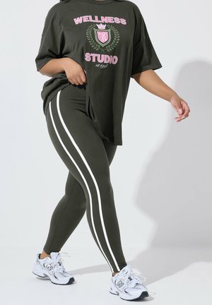 Person iført mørkegrønne leggings med hvide sidelommer og en matchende oversized "Wellness Studio" t-shirt, gående i hvide sneakers.