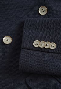 Tengerészkék textúrált szövet öt kerek gombbal; négy csoportosítva, kontrasztos bézs és fekete varrási részletekkel.