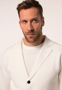 Cárdigan blanco de punto con botón negro, que presenta un cuello chal y una tela estructurada, superpuesto a una camiseta blanca sencilla.