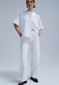 Persoon draagt een loszittend wit overhemd met korte mouwen en wijde witte broek, staat met de handen in de zakken tegen een effen achtergrond.