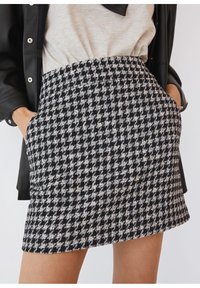 Femme portant une jupe à motif pied-de-poule noir et blanc avec poches latérales, un haut beige et une veste en cuir noire, mains dans les poches.